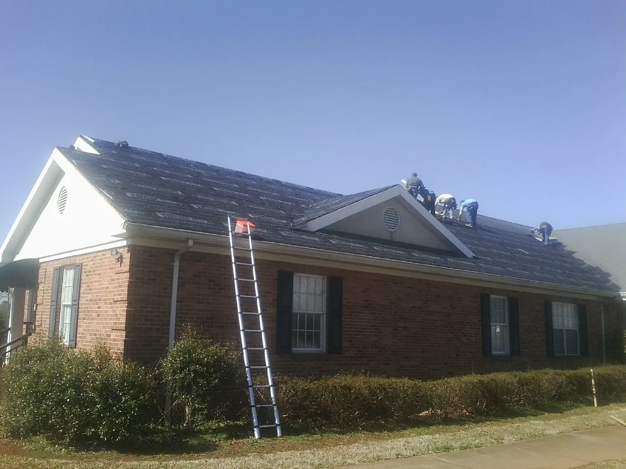 Crew tearing shingles off a brick ranch roof with a ladder set — Emergency Roof Repair work by our crew in Attica