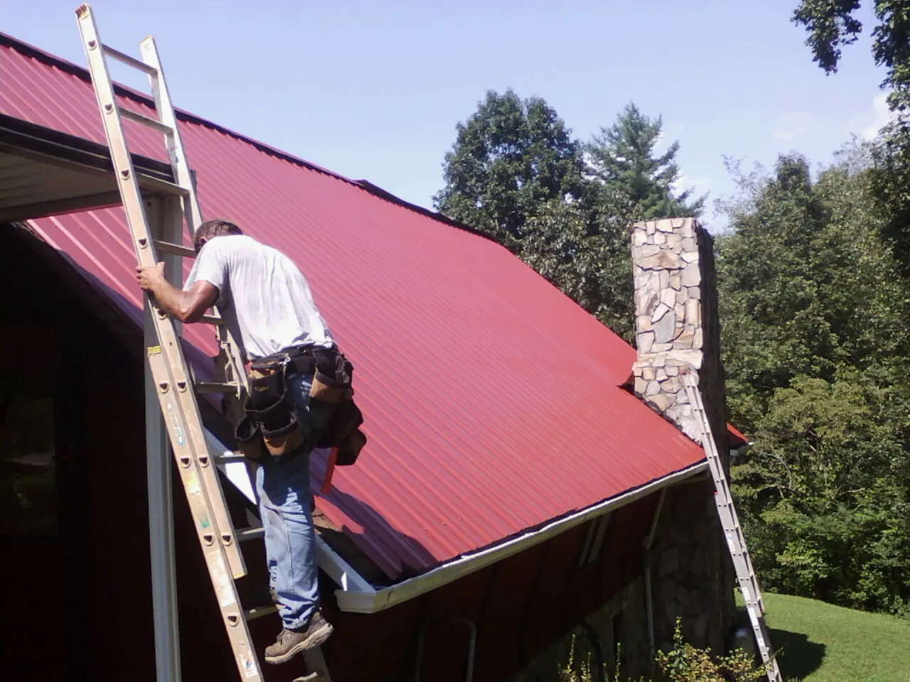 Roofer on a ladder beside a red metal roof and stone chimney — Soffit & Fascia Repair by Roofing Brothers in Attica
